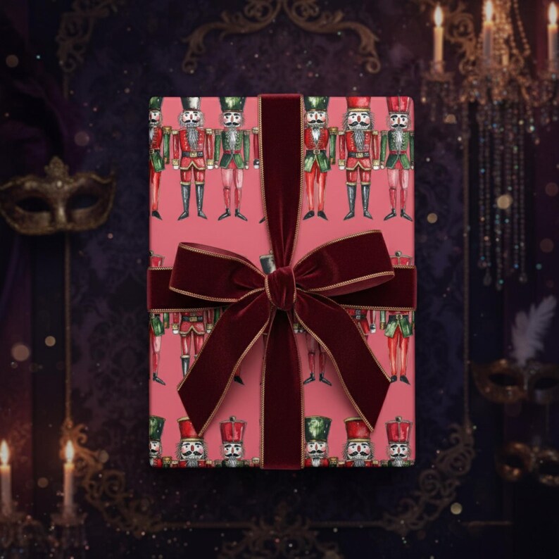 May include: A wrapped gift box featuring a pink background with a repeating pattern of nutcracker soldiers in red and green uniforms. The box is tied with a wide, dark red velvet ribbon and bow, creating a festive and elegant presentation.