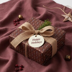 May include: A wrapped gift box with a brown patterned design, tied with a burlap ribbon and a tag that reads "Happy Holidays." The gift is decorated with a sprig of evergreen and sits on a maroon fabric surface.