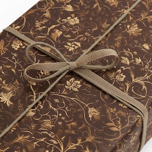 May include: A wrapped gift box with brown floral patterned paper and a tan ribbon tied in a bow. The paper has a gold floral design. The ribbon is wrapped around the box and tied in a bow on top.