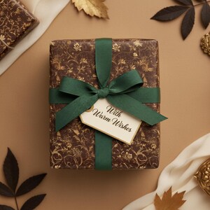 May include: A wrapped gift box with a brown floral pattern and a dark green ribbon and bow. A tag that says "With Warm Wishes" is attached to the bow. The gift is on a tan surface with decorative leaves.