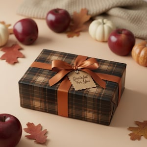Puede incluir: Una caja de regalo envuelta en papel a cuadros marr&oacute;n, atada con una cinta y un lazo color &oacute;xido. Una etiqueta que dice "Grateful For You" cuelga del lazo. La escena incluye manzanas rojas, peque&ntilde;as calabazas y hojas de oto&ntilde;o.