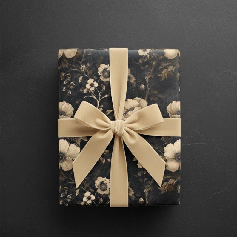 May include: A rectangular gift box wrapped in dark floral patterned paper, tied with a wide, light tan velvet ribbon and bow. The floral design features white and cream-colored flowers and foliage against a dark background.