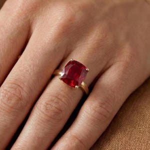 May include: A gold ring featuring a large, square-cut, deep red gemstone. The ring is set on a slender gold band, showcasing the vibrant color of the stone. The ring is displayed on a hand.
