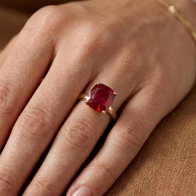 May include: A gold ring featuring a large, square-cut, deep red gemstone. The ring is set on a slender gold band, showcasing the vibrant color of the stone. The ring is displayed on a hand.