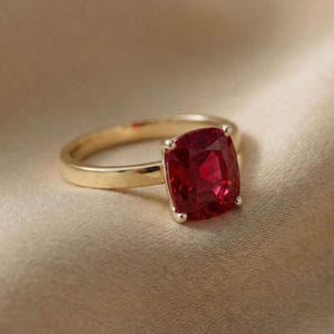 May include: A gold ring featuring a large, square-cut, deep red gemstone. The ring's band is a polished gold, and the gemstone is held in place by four small prongs. The ring is set against a neutral, textured background.