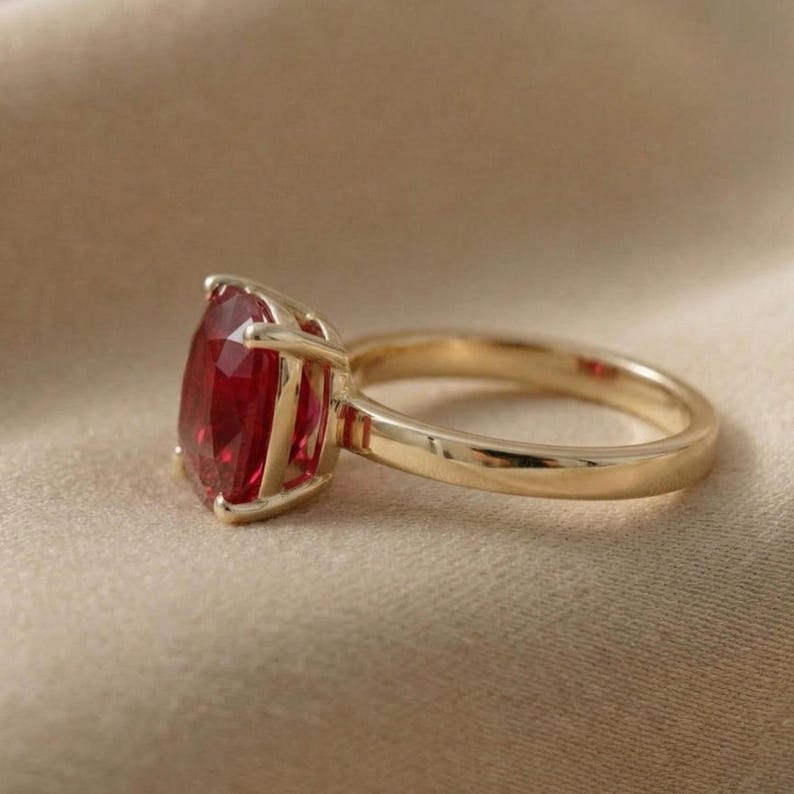 May include: A gold ring with a large, faceted, red gemstone. The gemstone is pear-shaped and set in a gold band. The ring is displayed on a neutral-colored surface.