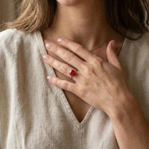 May include: A gold ring featuring a large, square-cut, vibrant red gemstone. The ring is set on a delicate gold band. The image shows the ring on a person's finger, with a gold necklace visible.