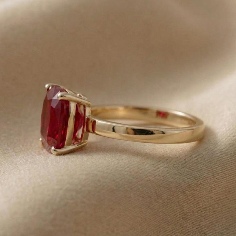 May include: A gold ring with a large, faceted, oval-shaped red gemstone. The gemstone is set in a four-prong setting. The ring band is smooth and polished, and the overall design is elegant and classic.