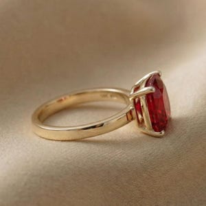 May include: A gold ring with a large, pear-shaped, red gemstone. The ring's band is smooth and polished, and the gemstone is held in place by four prongs. The ring is set against a neutral background.