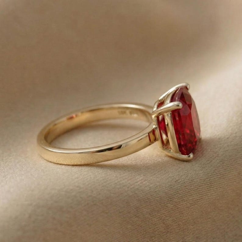 May include: A gold ring with a large, pear-shaped, red gemstone. The ring's band is smooth and polished, and the gemstone is held in place by four prongs. The ring is set against a neutral background.