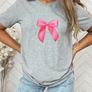 May include: A gray t-shirt with a pink bow printed on the front.