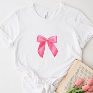 May include: White t-shirt with a pink bow graphic printed on the front. The shirt is laying flat on a white surface with a book and flowers next to it.