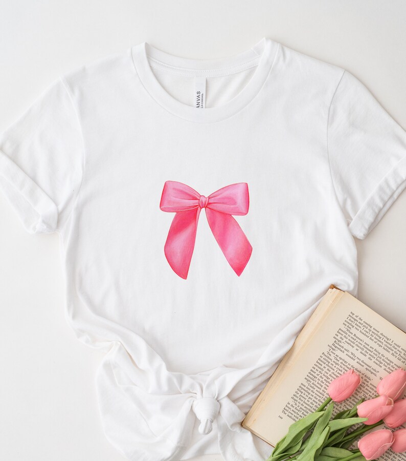 May include: White t-shirt with a pink bow graphic printed on the front. The shirt is laying flat on a white surface with a book and flowers next to it.