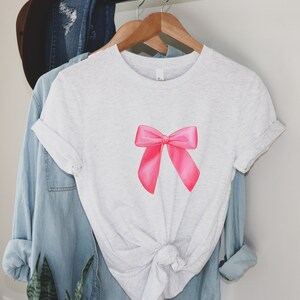 May include: A white t-shirt with a pink bow on the front. The shirt is knotted at the bottom.