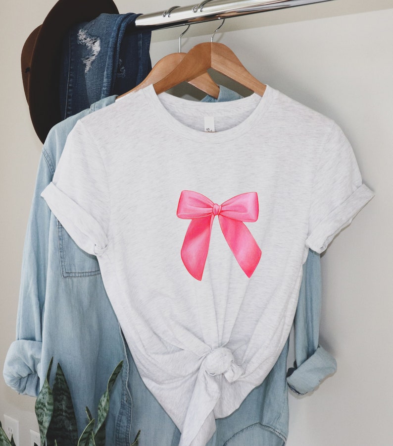 May include: A white t-shirt with a pink bow on the front. The shirt is knotted at the bottom.
