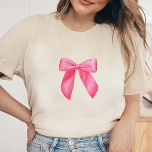 May include: A cream colored t-shirt with a pink bow design on the front.