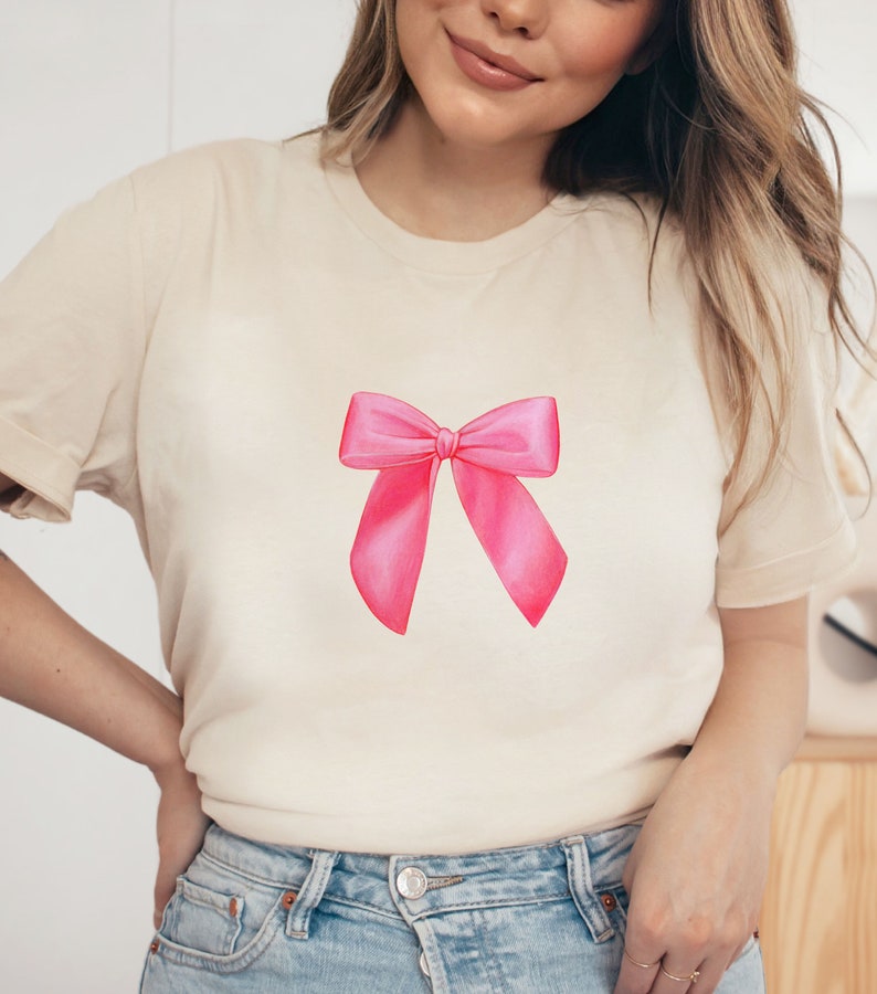 May include: A cream colored t-shirt with a pink bow design on the front.