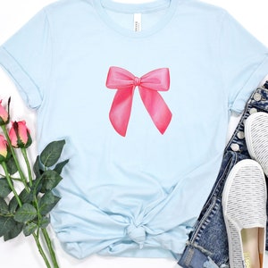 May include: A light blue t-shirt with a pink bow design. The shirt is made of a soft, comfortable material and has a relaxed fit.