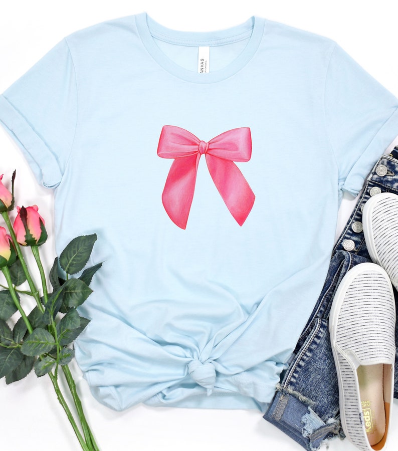 May include: A light blue t-shirt with a pink bow design. The shirt is made of a soft, comfortable material and has a relaxed fit.