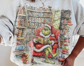May include: A light gray sweatshirt featuring a colorful illustration of a green character in a Santa suit reading a book in a red armchair. The background depicts a library with bookshelves and presents.