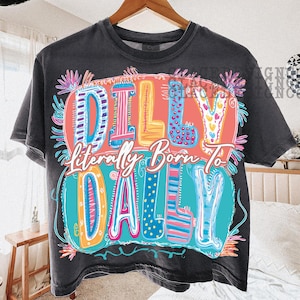 May include: Dark gray t-shirt with the text "Dilly Literally Born To Dally" in colorful, bubble-style letters. The design includes pink, blue, yellow, and orange accents, and is surrounded by floral elements. The shirt is hanging on a wooden hanger.