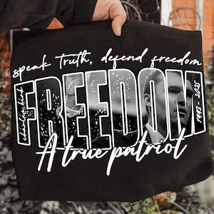 May include: Black tote bag featuring the word "FREEDOM" in white, with a black and white image of a man within the letters. The bag also has the text "speak truth, defend freedom" and "A true patriot".