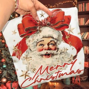 May include: White Christmas pillow featuring a vintage illustration of Santa Claus with a large red bow and gold stars. The words "Merry Christmas" are written in red script across the bottom. A festive holiday decoration.