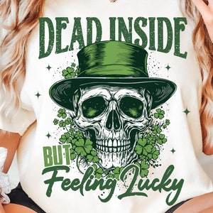 Op de afbeelding: Wit T-shirt met een groene schedel die een groene hoge hoed draagt en omringd is door klavertjesvier. De tekst "Dead Inside But Feeling Lucky" is op het shirt gedrukt.