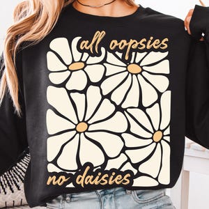 May include: Black sweatshirt with a white daisy print and the text "all oopsies no daisies" in yellow.