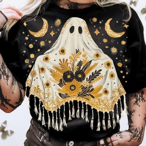 May include: Black t-shirt featuring a ghost design with a floral shawl. The ghost has two eye holes and is adorned with gold moons, stars, and floral patterns. The shawl has a fringe trim. The shirt is a crew neck.