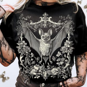 May include: Black t-shirt featuring a detailed grayscale illustration of a bat with outstretched wings, surrounded by floral elements and ornate scrollwork. The bat has a smiling expression. The shirt is a crew neck.