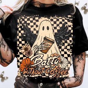 Peut inclure: T-shirt noir avec un motif de fantôme tenant une tasse de café, avec le texte "Batty Without Coffee". Le design comprend un fond à carreaux, des ailes et des motifs floraux orange, crème et noirs.