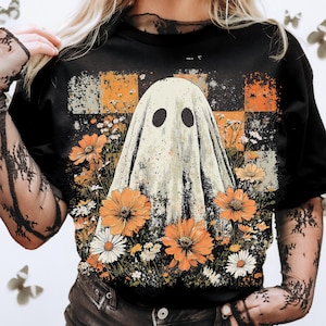 Puede incluir: Camiseta negra con un dise&ntilde;o de fantasma vintage, flores naranjas y blancas y un fondo a cuadros. El fantasma tiene dos agujeros negros para los ojos y est&aacute; rodeado de un arreglo floral. El fondo tiene un patr&oacute;n a cuadros en naranja, blanco y gris.
