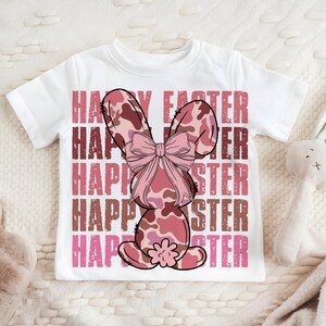 May include: White t-shirt featuring a pink and brown camouflage bunny rabbit wearing a pink bow. The shirt has the text "Happy Easter" repeated five times in pink and brown.