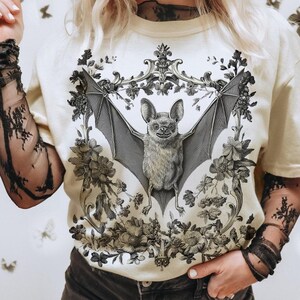 May include: Cream-colored t-shirt featuring a detailed black and white illustration of a bat with outstretched wings, surrounded by floral elements. The bat design is centered within an ornate frame, creating a gothic and botanical aesthetic.