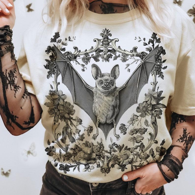 May include: Cream-colored t-shirt featuring a detailed black and white illustration of a bat with outstretched wings, surrounded by floral elements. The bat design is centered within an ornate frame, creating a gothic and botanical aesthetic.