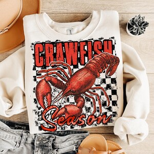 May include: A cream-colored sweatshirt with a red crawfish graphic and the text "Crawfish Season" in red letters. The crawfish is on a black and white checkered background.