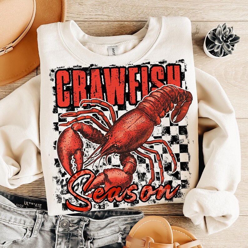 May include: A cream-colored sweatshirt with a red crawfish graphic and the text "Crawfish Season" in red letters. The crawfish is on a black and white checkered background.