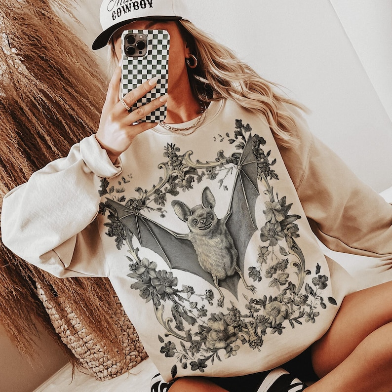 May include: Beige sweatshirt featuring a detailed illustration of a bat with outstretched wings, surrounded by floral elements. The bat design is in shades of gray and beige. The sweatshirt has long sleeves and a crew neck.