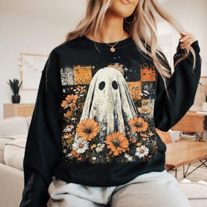 Puede incluir: Sudadera negra con un dise&ntilde;o de fantasma vintage, rodeada de flores naranjas y blancas. El fantasma tiene dos ojos negros. El fondo presenta un patr&oacute;n de cuadros en naranja, blanco y gris.
