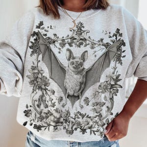 May include: Light gray sweatshirt featuring a detailed illustration of a bat with outstretched wings, surrounded by floral and ornamental elements. The bat is the focal point, rendered in grayscale, with intricate details. The design is centered on the front of the sweatshirt.
