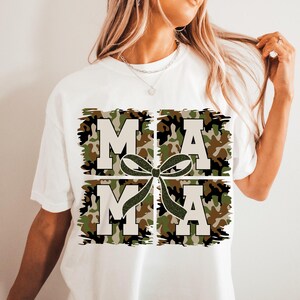Pu&ograve; includere: Maglietta bianca con un design a stampa mimetica che scrive "MAMA" con un fiocco verde al centro.