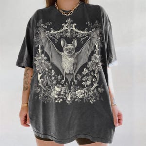 May include: Dark gray t-shirt featuring a detailed illustration of a bat with outstretched wings, surrounded by floral elements. The bat is centered within an ornate frame, creating a gothic and vintage aesthetic. The shirt has a slightly faded appearance.