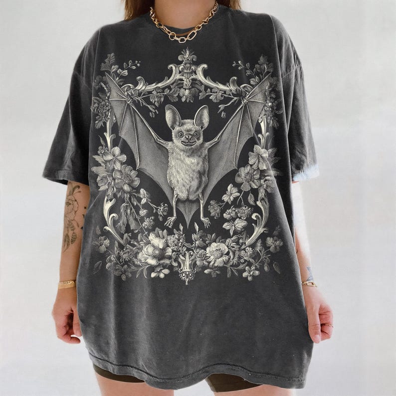 May include: Dark gray t-shirt featuring a detailed illustration of a bat with outstretched wings, surrounded by floral elements. The bat is centered within an ornate frame, creating a gothic and vintage aesthetic. The shirt has a slightly faded appearance.
