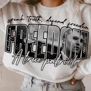 May include: A light grey sweatshirt featuring the word "FREEDOM" in large, bold, black and white letters. The word "FREEDOM" has a black and white image of a man inside. The sweatshirt also has the phrases "speak truth, defend freedom", "A true patriot", and "1993-2025" printed on it.