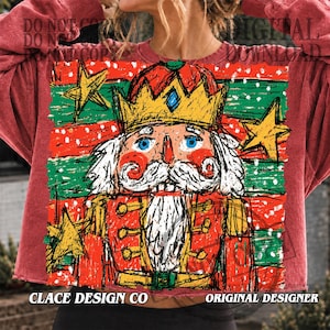 May include: A red crewneck sweatshirt featuring a graphic print of a nutcracker wearing a gold crown, with a white moustache and beard. The background consists of horizontal red and green stripes with yellow stars and white snow-like dots.