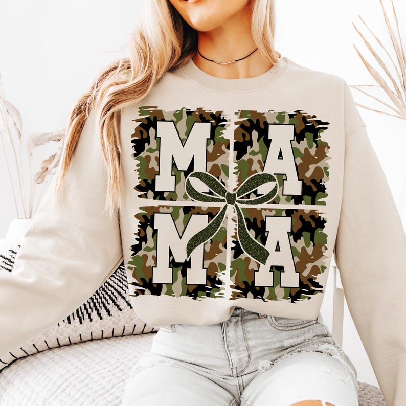 Pu&ograve; includere: Una felpa beige con un design grafico della parola "MAMA" in lettere bianche con un motivo mimetico verde, marrone e nero. Le lettere sono in una scatola con un fiocco verde e nero.
