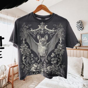 May include: Dark gray t-shirt featuring a detailed illustration of a bat with outstretched wings, surrounded by floral and ornamental elements. The bat is the central focus, with intricate shading and texture. The shirt is hanging on a wooden hanger.