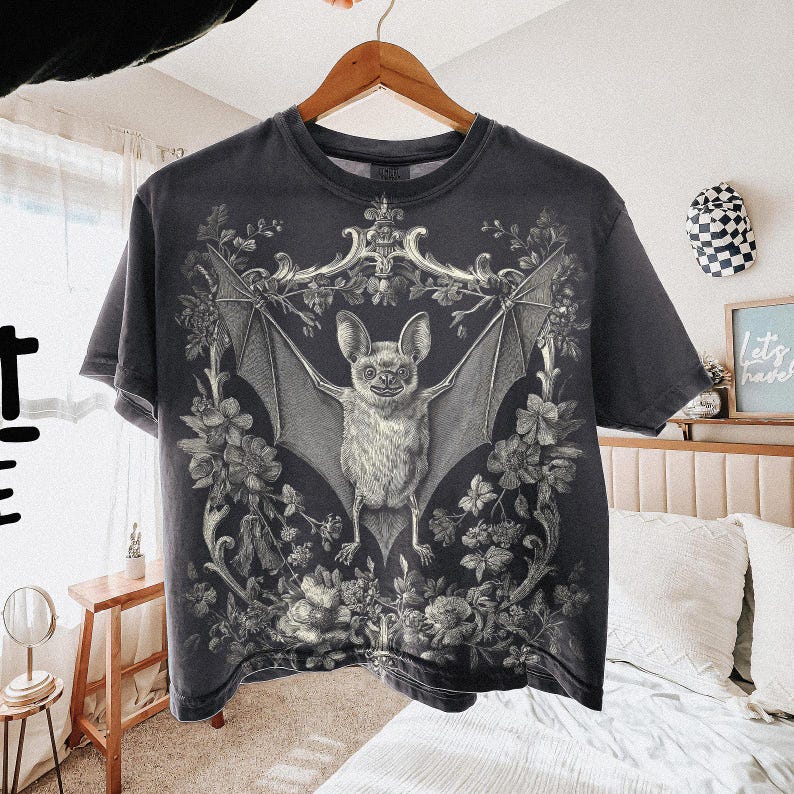 May include: Dark gray t-shirt featuring a detailed illustration of a bat with outstretched wings, surrounded by floral and ornamental elements. The bat is the central focus, with intricate shading and texture. The shirt is hanging on a wooden hanger.