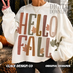 May include: A cream-colored long-sleeve sweatshirt featuring the text HELLO FALL in large, multi-colored block letters. The design includes small, scattered autumn leaf illustrations in shades of orange, brown, and green, printed on the front.